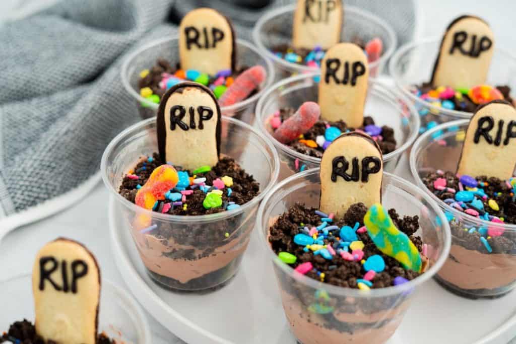A tray of Halloween graveyard dirt cups made with chocolate pudding and crushed cookies, each topped with a cookie tombstone labeled “RIP” in black icing, colorful spooky sprinkles, and gummy worms. The treats are arranged in clear plastic cups for a festive, kid-friendly dessert display.