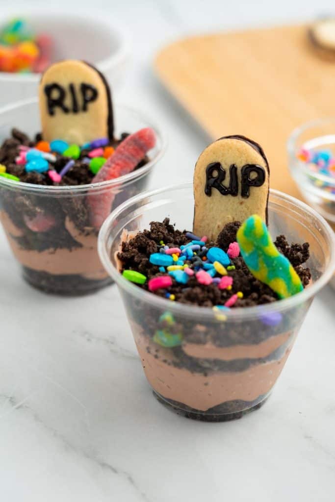 Close-up of two Halloween dirt and worms pudding cups layered with chocolate pudding and crushed cookies, topped with Milano cookie tombstones labeled “RIP,” colorful sprinkles, and gummy worms for a spooky dessert effect.
