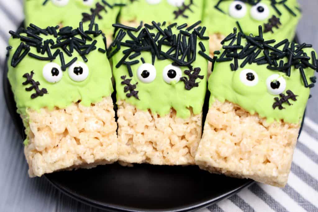 Frankenstein monster Rice Krispie treats lined up on a black plate, each decorated with green frosting, candy eyes, black sprinkle hair, and chocolate stitch details to resemble the iconic Halloween character. The treats rest on a gray and white striped cloth.
