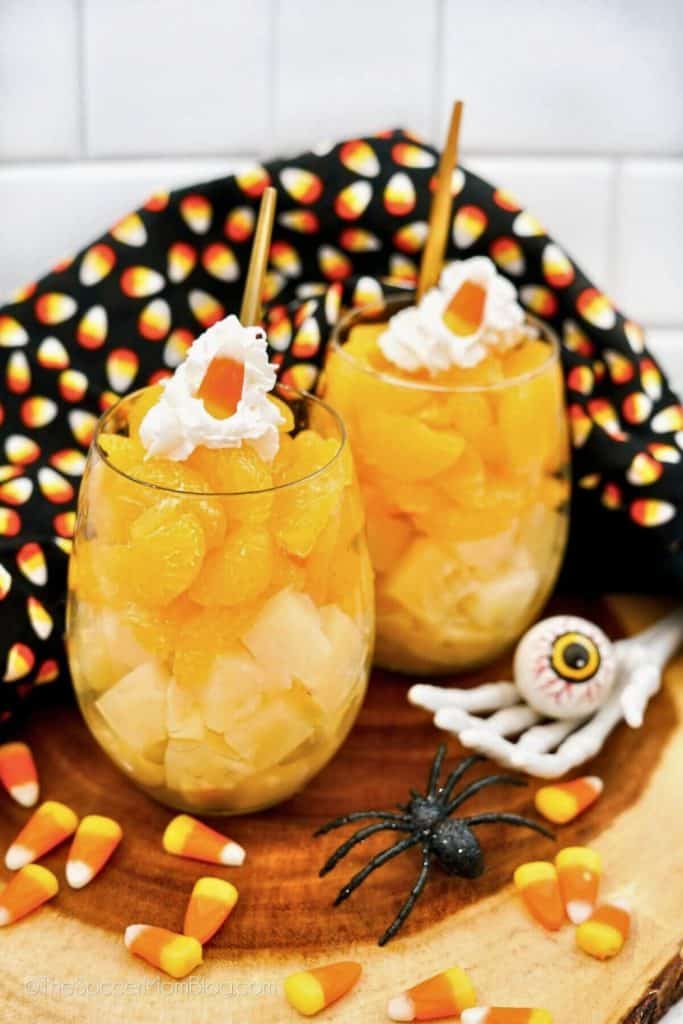 Two layered fruit cups made with pineapple chunks and mandarin oranges, topped with whipped cream and a single candy corn, served in stemless glasses with gold spoons. Halloween-themed setting includes candy corn scattered on a wooden surface, a plastic spider, and a skeleton hand holding an eyeball.