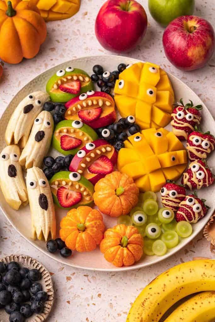 Plate of healthy no bake Halloween treats made from fresh fruit, including banana ghosts, apple monster mouths with sunflower seed teeth and strawberry tongues, mandarin orange pumpkins, mango cubes with candy eyes, and strawberry mummies drizzled with white chocolate. Fun and colorful Halloween snack display perfect for kids.