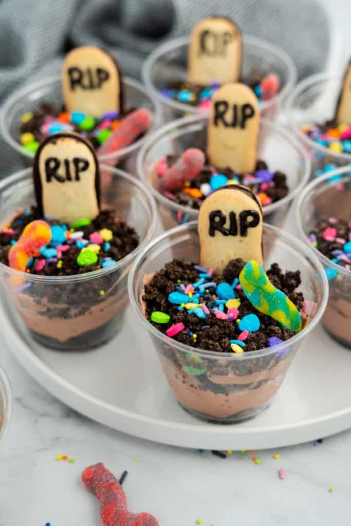 Tray of Halloween graveyard pudding cups featuring layers of chocolate pudding and crushed oreo cookies, each topped with a Milano cookie tombstone labeled “RIP,” colorful spooky sprinkles, and gummy worms. The playful dessert is designed to look like a mini graveyard scene.