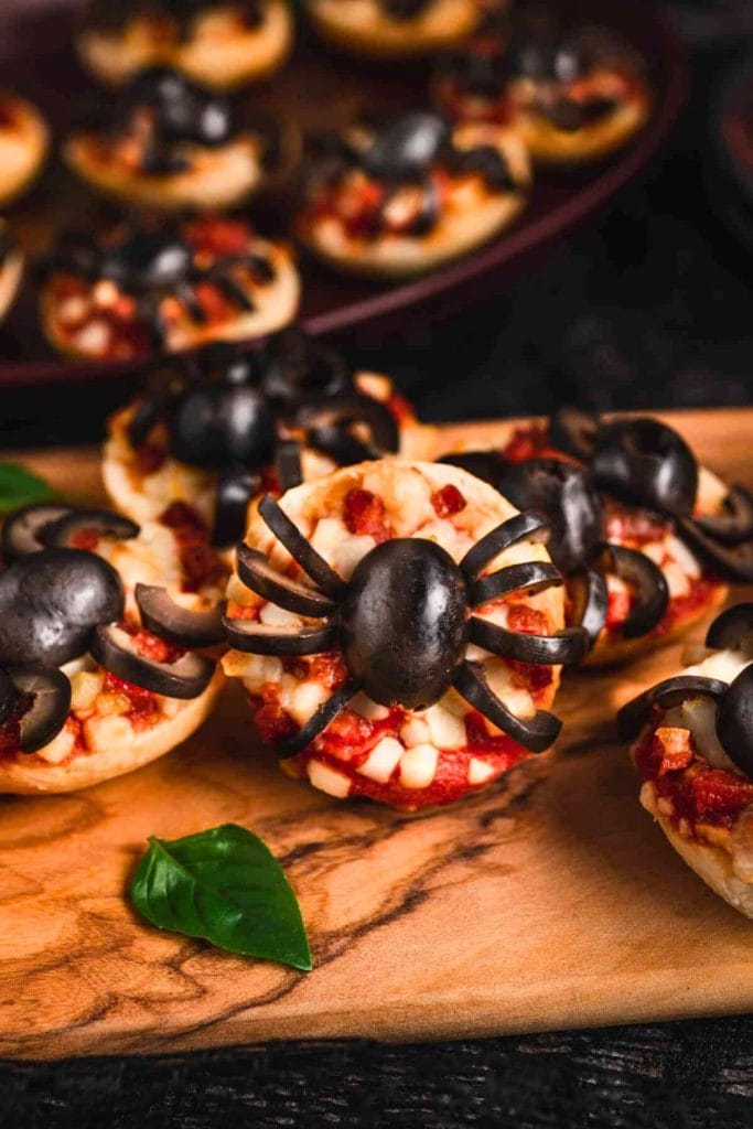 Close-up of Halloween mini spider pizzas topped with black olive spiders on melted cheese and tomato sauce, arranged on a wooden serving board with a fresh basil leaf for garnish. More mini pizzas are visible in the background on a dark tray, creating a festive and spooky snack display.