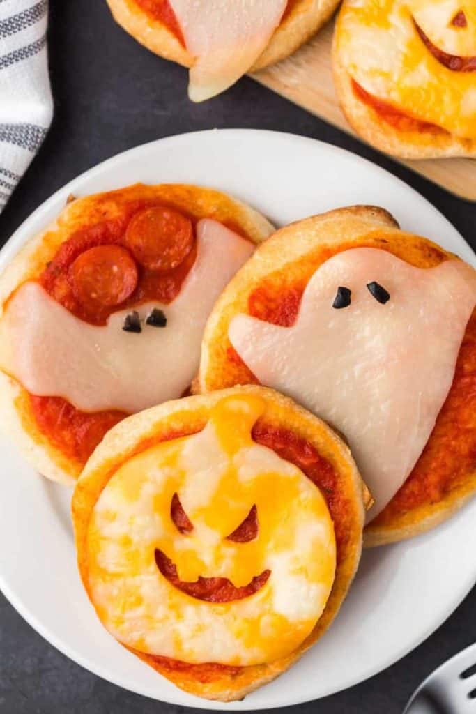 Plate of Halloween Mini Pizzas topped with festive shapes made from cheese, including a jack-o'-lantern, a ghost, and a bat with pepperoni eyes. Each pizza features a golden crust, tomato sauce, and playful Halloween-themed toppings perfect for a spooky snack.