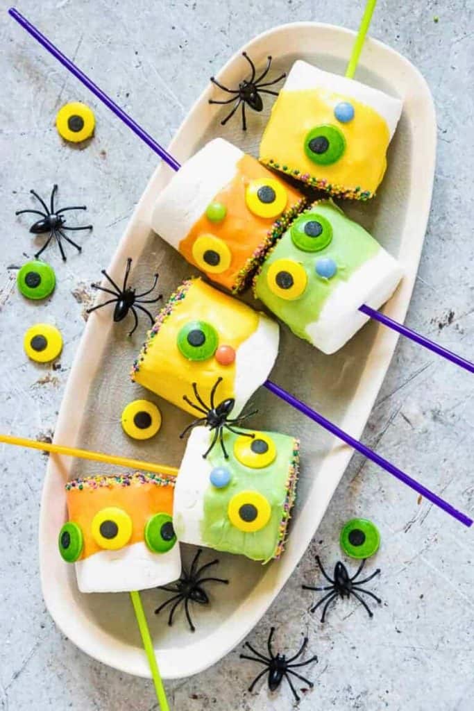 Colorful Halloween marshmallow pops dipped in yellow, orange, and green candy coating, decorated with candy eyes and rainbow sprinkles, each on a colorful stick. Displayed on a white tray with plastic spiders and extra candy eyes scattered around.