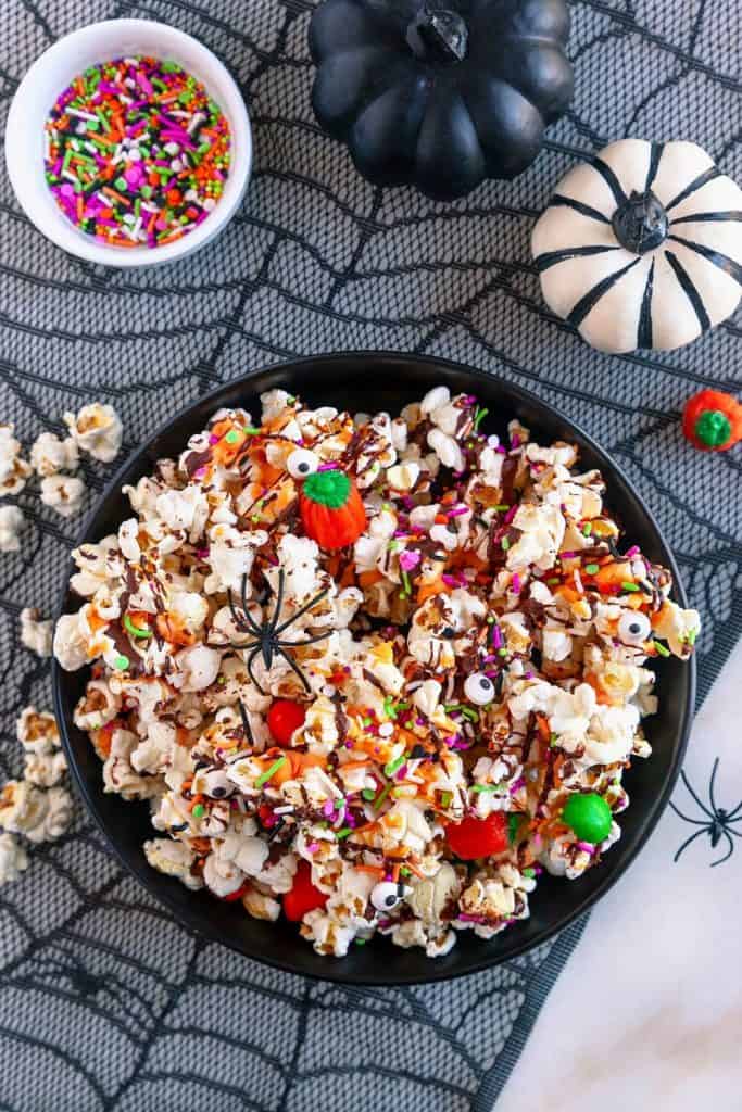 Bowl of Halloween popcorn mix topped with candy eyeballs, chocolate drizzle, orange icing, colorful sprinkles, candy pumpkins, and red and green candies. Set on a spiderweb tablecloth with mini pumpkins and spooky decorations for a festive and fun Halloween treat.
