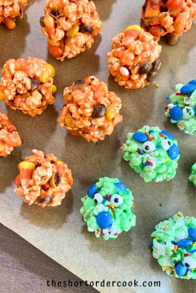 Halloween popcorn balls in festive colors, with orange popcorn clusters mixed with candy corn and chocolate candies, and green monster-themed popcorn balls decorated with candy eyes, blue M&Ms, and rainbow sprinkles. Arranged on a parchment-lined surface.