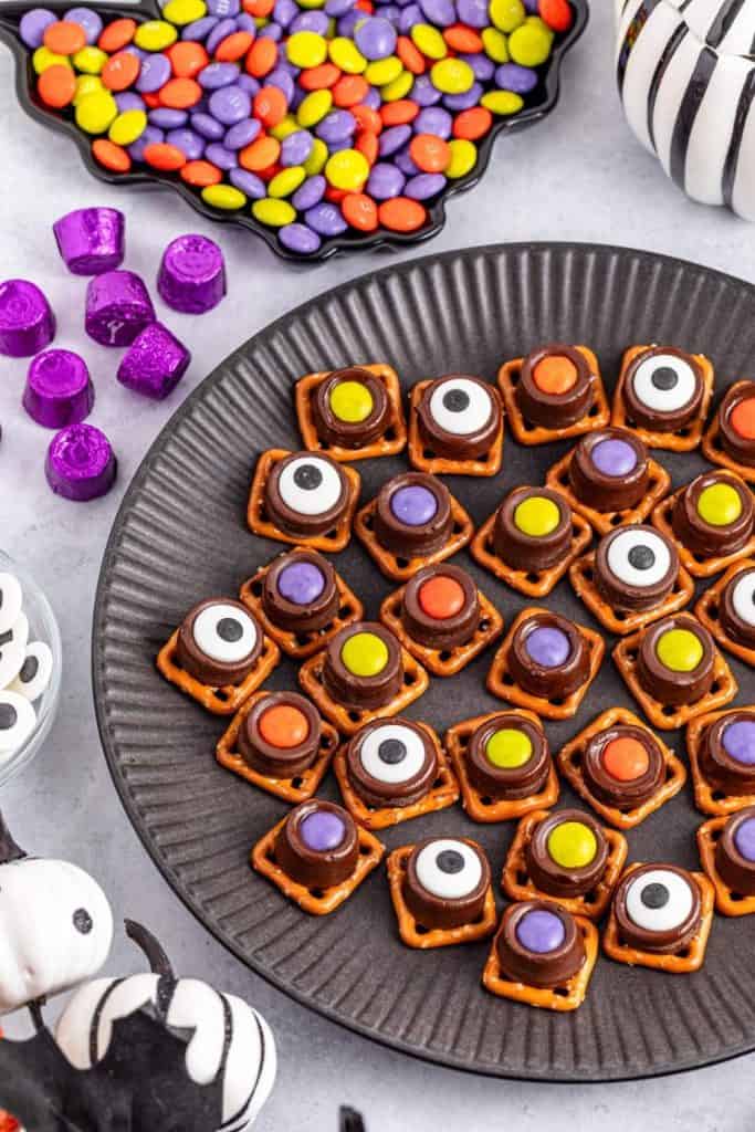 Round black plate filled with Halloween pretzel snacks made by topping square pretzels with melted chocolate candies and colorful Halloween-themed candy eyes and M&Ms. Surrounded by purple-wrapped chocolates, candy eyes, and festive Halloween decorations for a playful and spooky treat display.