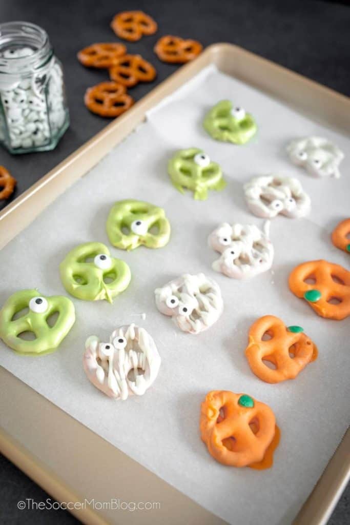 Tray of Halloween-themed pretzels dipped in colorful candy coating, including green pretzels with candy eyes, white mummy pretzels with drizzled chocolate, and orange pumpkin pretzels with green candy stems. Displayed on parchment paper with plain pretzels and a jar of candy eyes in the background.