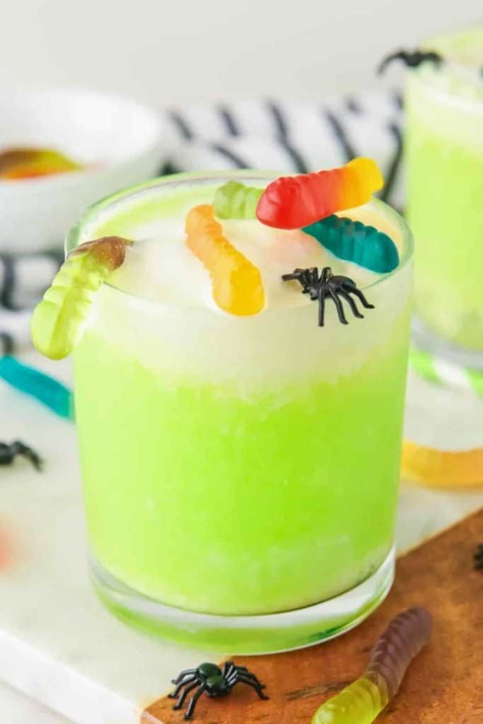 Bright green Halloween drink served in a glass and topped with whipped cream, colorful gummy worms, and small plastic spiders for a spooky garnish. The creepy-crawly themed beverage sits on a white tray with extra gummies and spiders scattered around.