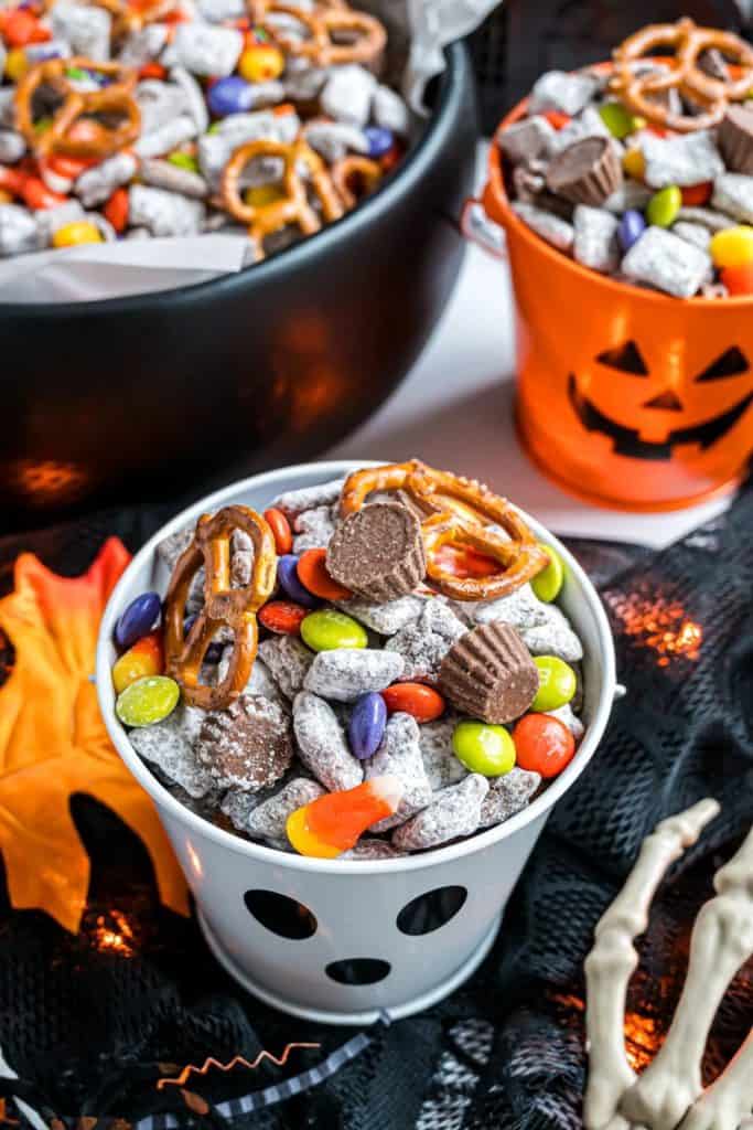 Halloween snack mix served in festive ghost and pumpkin buckets, filled with powdered puppy chow, mini peanut butter cups, candy-coated chocolates, candy corn, and pretzels. The treats are displayed on a black mesh cloth with fall leaves and skeleton props for a spooky movie night vibe.