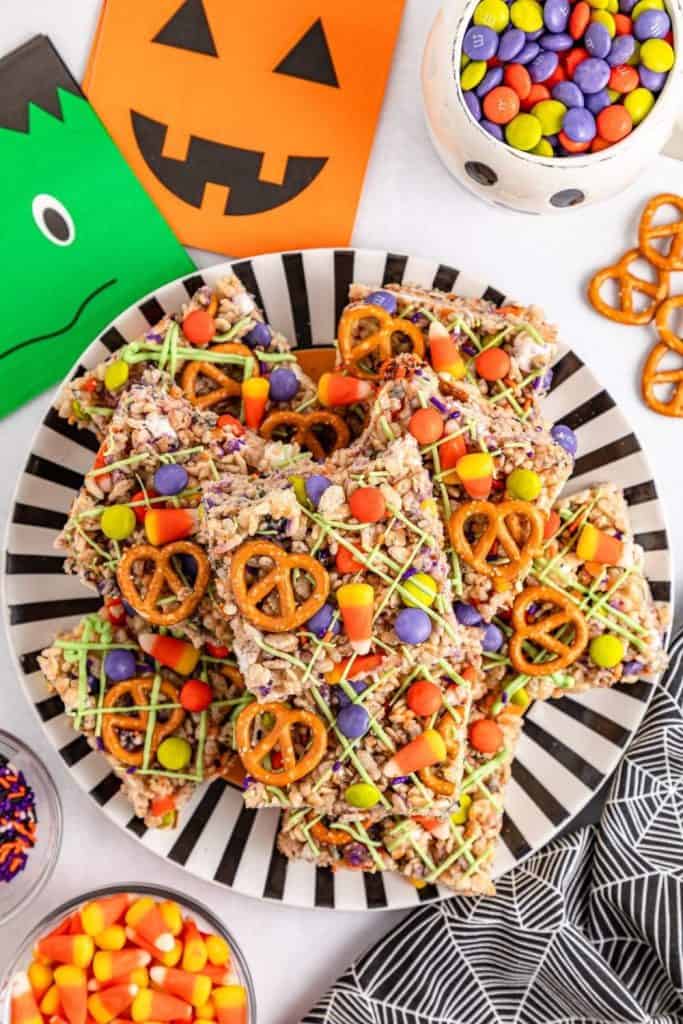 Festive Halloween Rice Krispie treats topped with pretzels, candy corn, colorful M&M-style candies, and green drizzle, served on a black-and-white striped plate. Surrounded by Halloween-themed napkins, candy dishes, and a spiderweb-patterned cloth for a playful spooky vibe.