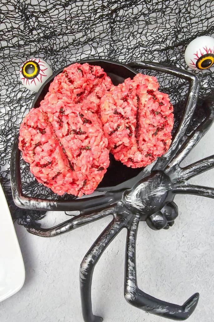 Three bright pink rice krispie treats shaped like brains with dark chocolate vein details, displayed on a black spider-shaped serving platter with fake eyeballs and a spooky net backdrop.