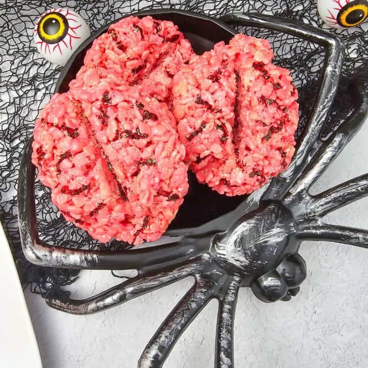Three bright pink rice krispie treats shaped like brains with dark chocolate vein details, displayed on a black spider-shaped serving platter with fake eyeballs and a spooky net backdrop.