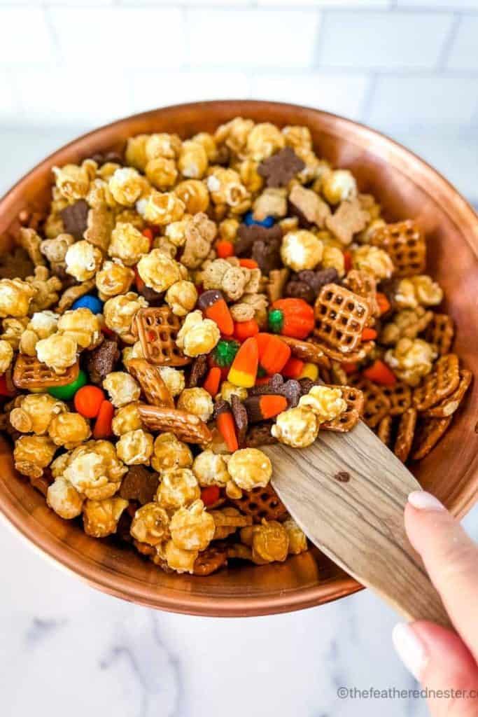 Large wooden bowl filled with Halloween snack mix featuring caramel popcorn, mini pretzels, candy corn, pumpkin candies, chocolate cereal pieces, and colorful M&M-style chocolates. A festive sweet and salty blend being scooped with a wooden spatula, perfect for Halloween parties.