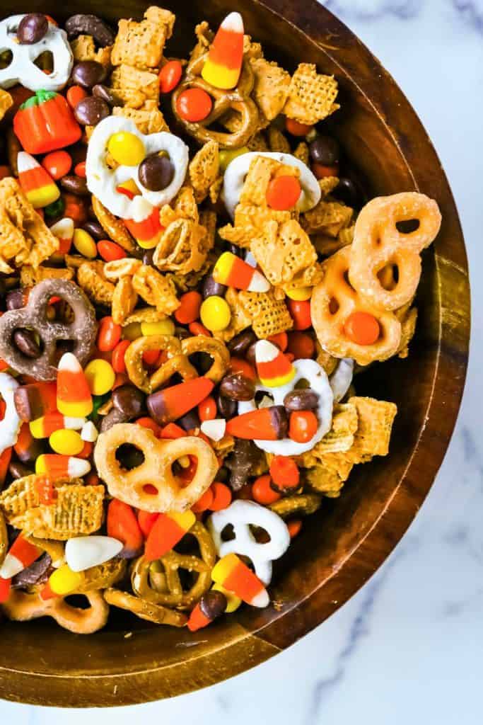 Wooden bowl filled with Halloween Snack Mix, including candy corn, candy pumpkins, chocolate-covered pretzels, Reese’s Pieces, Chex cereal, and yogurt-dipped pretzels. The mix is brightly colored in festive orange, yellow, brown, and white for a fun and crunchy Halloween treat.
