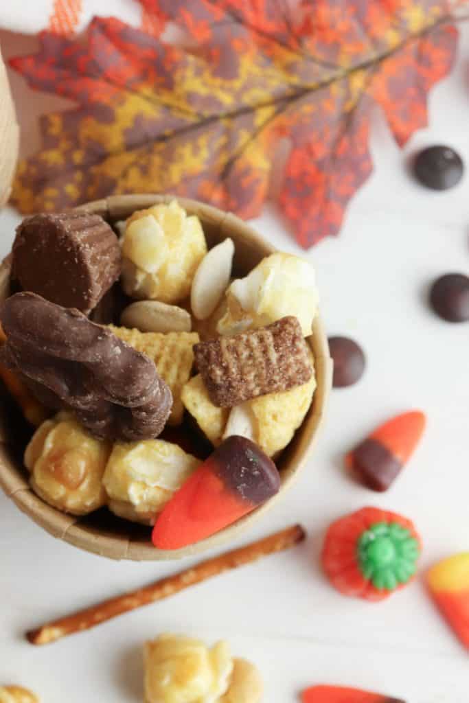 Small brown cup filled with Halloween Chex mix party snack, including chocolate-covered pretzel, caramel popcorn, Chex cereal, Bugles, candy corn, mini peanut butter cup, and pumpkin seeds, with scattered fall-themed candy and decorations on a white surface.