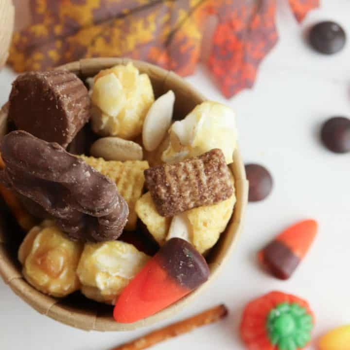 Small brown cup filled with Halloween Chex mix party snack, including chocolate-covered pretzel, caramel popcorn, Chex cereal, Bugles, candy corn, mini peanut butter cup, and pumpkin seeds, with scattered fall-themed candy and decorations on a white surface.