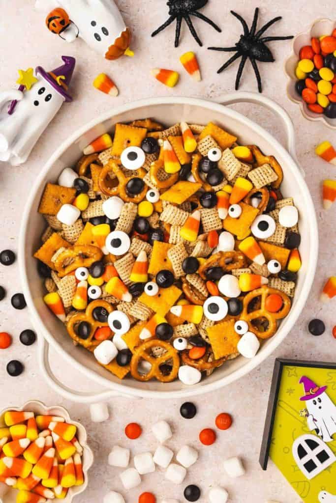 Large white bowl filled with Halloween snack mix made of Chex cereal, mini pretzels, Cheez-Its, candy corn, marshmallows, candy eyes, and chocolate candies in festive colors. Surrounded by Halloween-themed decorations like black spiders, ghost figurines, and small bowls of candy for a playful, spooky setup.