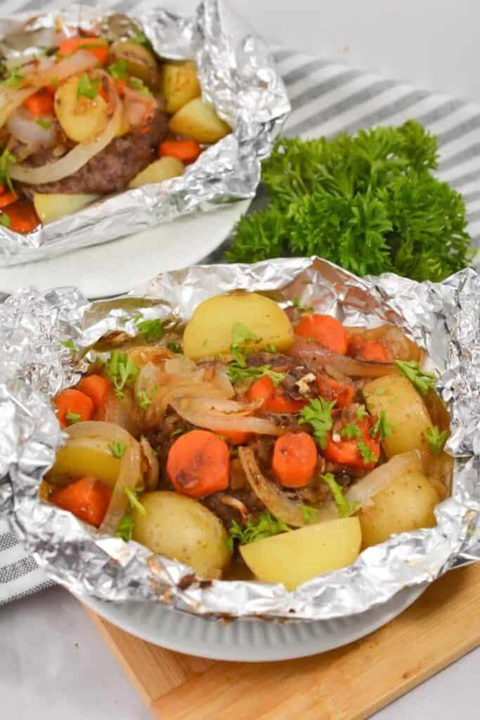 A plate topped with a round tin foil pack of hambuger patty, potatoes, carrots and onions. 