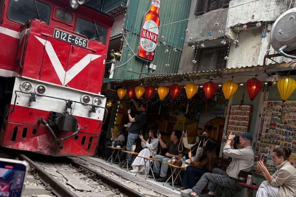 A red and white locomotive passes just inches from seated tourists at a narrow café-lined stretch of Hanoi Train Street. Colorful lanterns hang overhead as visitors eagerly snap photos and videos of the train from their sidewalk seats.