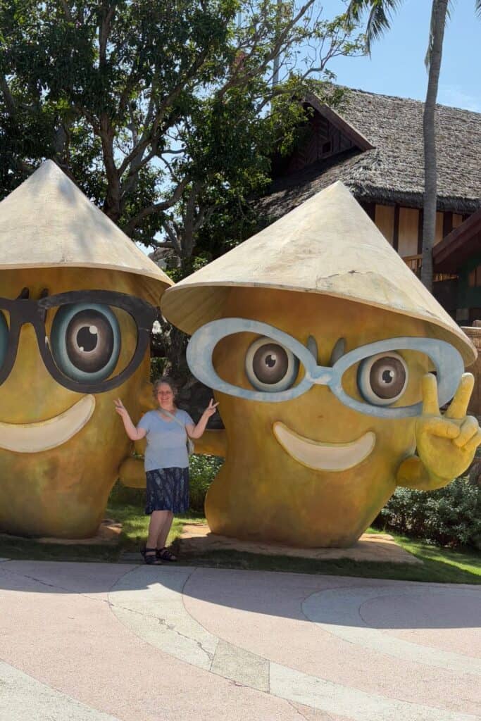 A woman poses with a peace sign between two large, cartoonish yellow mascots wearing oversized glasses and Vietnamese conical hats at Sun World’s Hon Thom Park in Vietnam. The cheerful statues smile widely, and lush greenery and a thatched-roof building create a tropical backdrop.