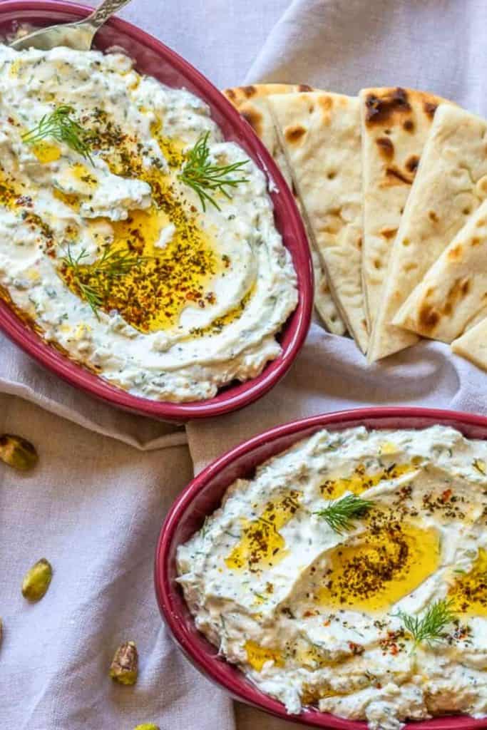 Creamy Haydari Turkish Yoghurt Dip served in red oval dishes, garnished with olive oil, dried herbs, and fresh dill. Presented alongside warm, sliced flatbread on a light cloth, with pistachios scattered nearby.