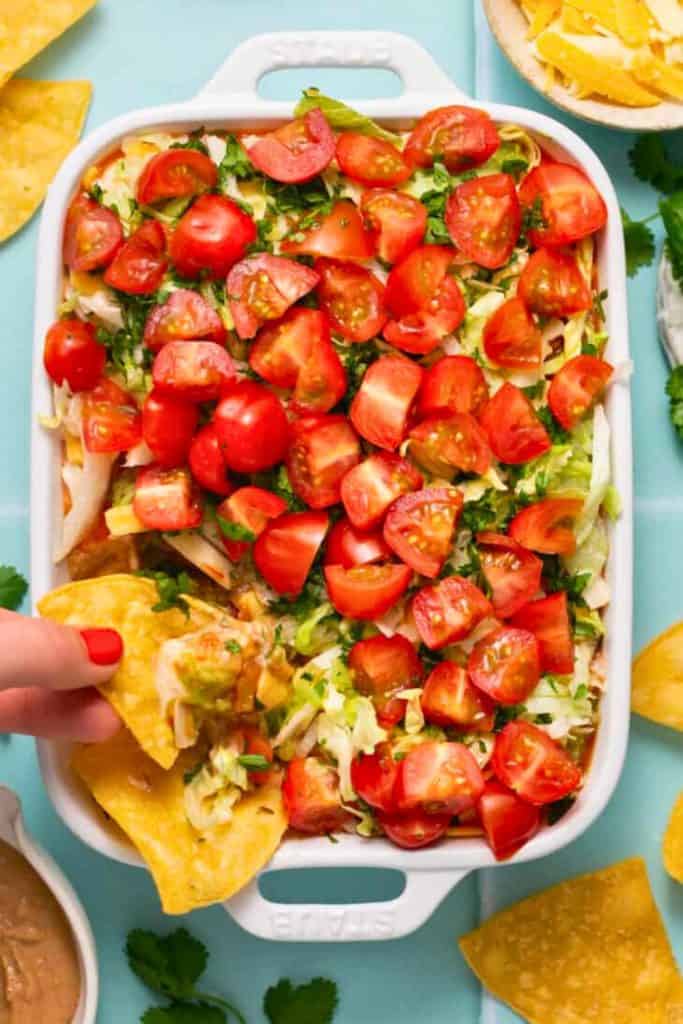 A vibrant healthy 7 layer dip in a white baking dish, topped with halved cherry tomatoes, shredded lettuce, and chopped cilantro. A hand with red nail polish dips a tortilla chip into the colorful layers, revealing creamy guacamole and beans underneath.