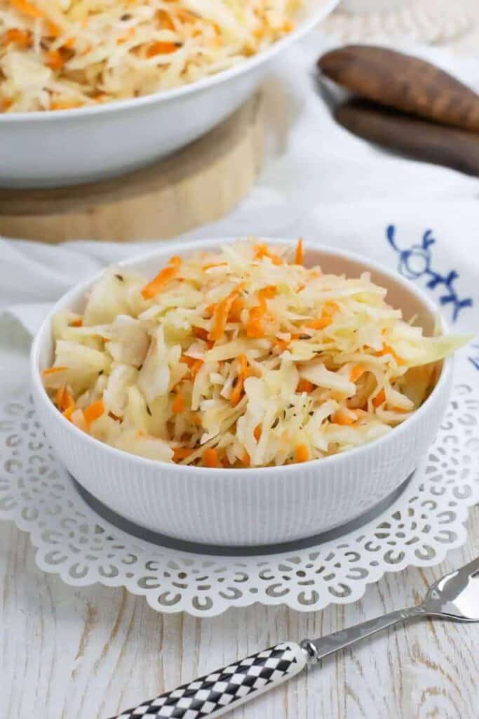 A white bowl filled with a light, vinegar-based apple coleslaw made with shredded cabbage, carrots, and apple slices, garnished with caraway seeds. The slaw sits on a white doily, with a patterned fork nearby.