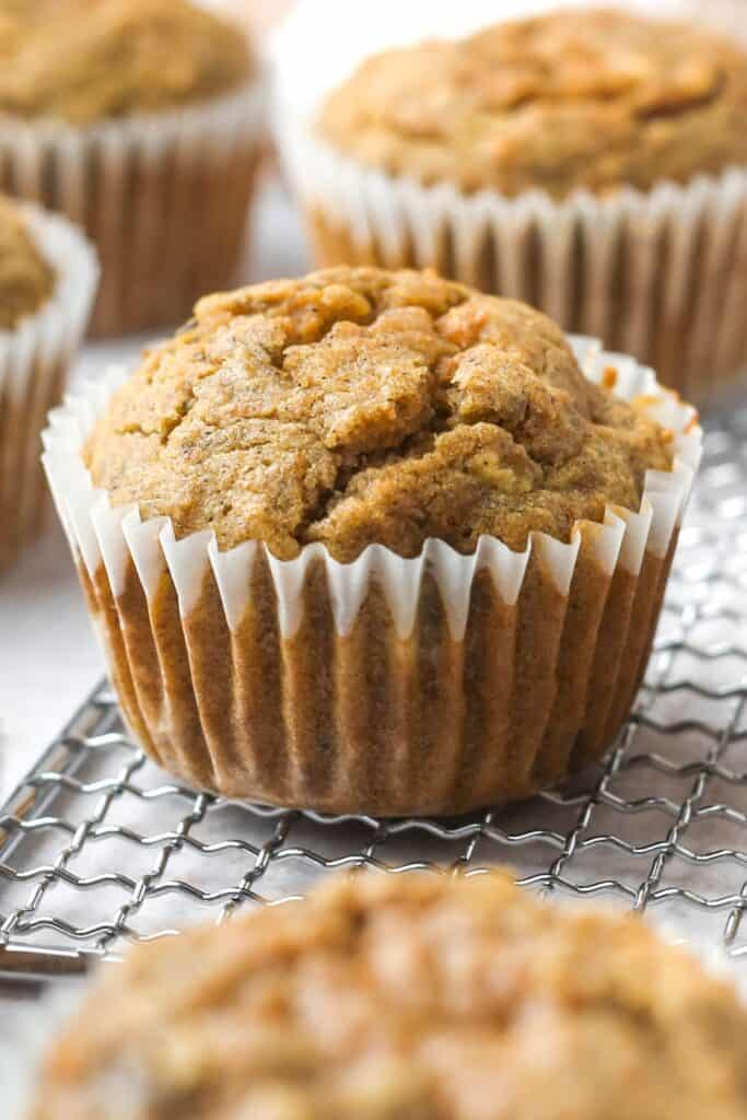 Soft golden brown banana carrot muffins in white paper liners cooling on a wire rack with a moist crumb and slightly cracked tops. These healthy banana carrot muffins are shown as a simple breakfast recipe ready to serve.
