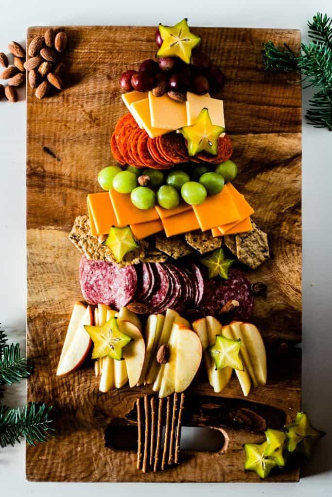 Wooden board with a Christmas tree made out of charcuterie including deli meats, red apples and green grapes. 