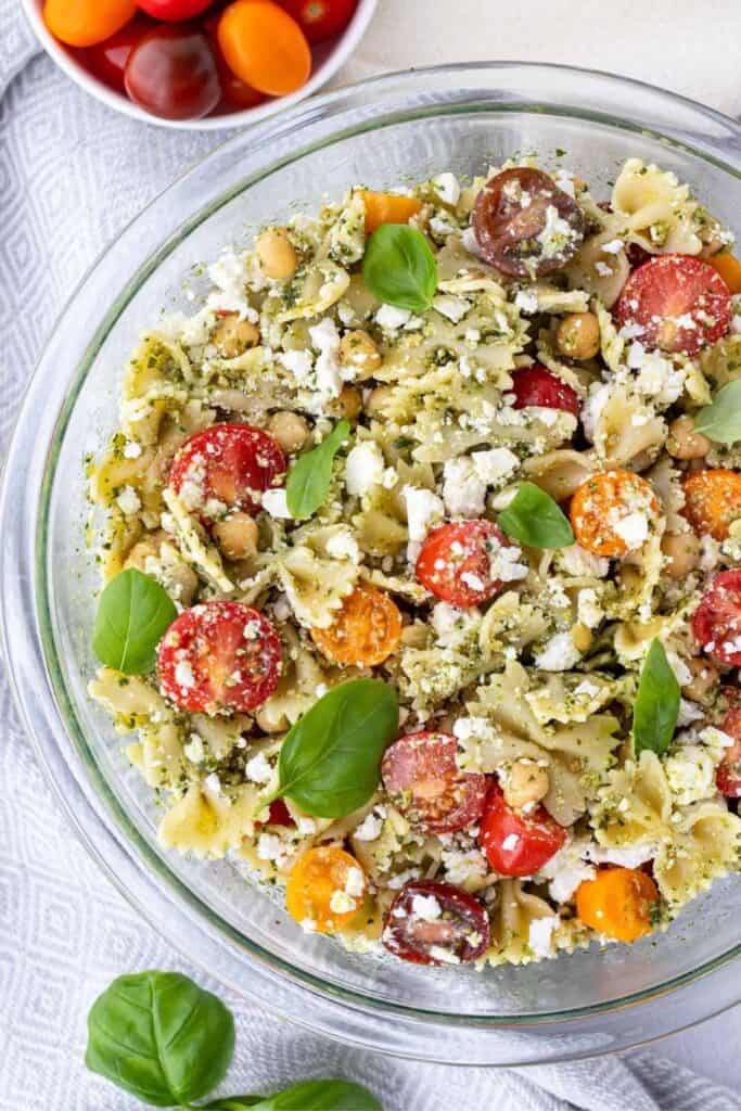 Large clear boel of healthy pesto, chickpea bow tie pasta salad with bell peppers, red and orange cherry tomatoes and feta cheese. 