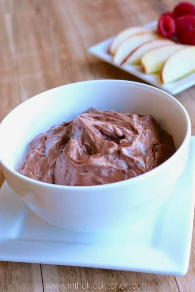 Smooth and creamy Healthy Chocolate Fruit Dip in a white bowl, set on a square white plate. A tray of fresh apple slices and raspberries is blurred in the background.