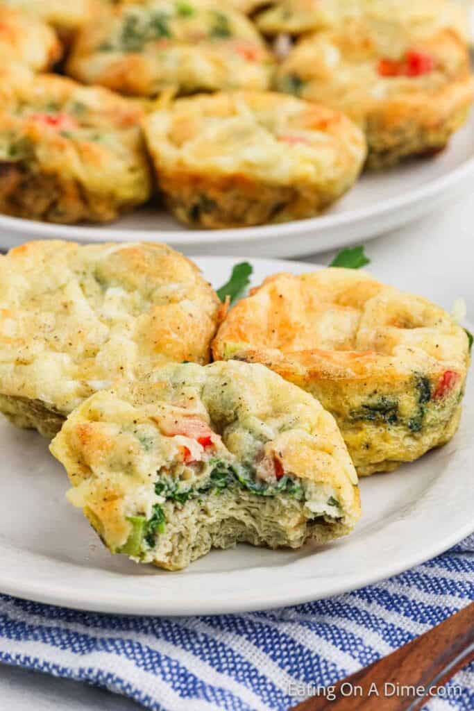 Close-up of three fluffy egg muffins filled with spinach, red bell pepper, and cheese, with one muffin showing a bite revealing the veggie-packed center. Served on a white plate with more muffins in the background, these savory egg bites make a healthy, grab-and-go breakfast or lunch option.