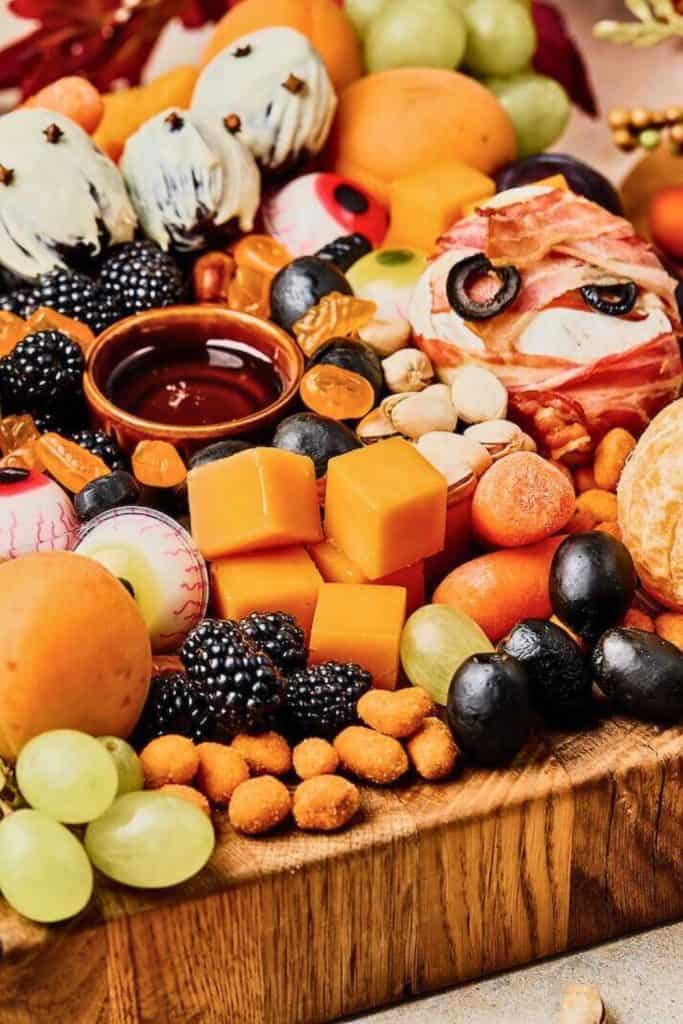 Healthy Halloween snack board for movie nights featuring a festive spread of fruits, cheese cubes, nuts, blackberries, olives, and themed treats like ghost-dipped strawberries, candy eyeballs, and a bacon-wrapped cheese ball decorated as a mummy. A small bowl of dip sits in the center, surrounded by colorful and spooky snacks on a wooden serving board.