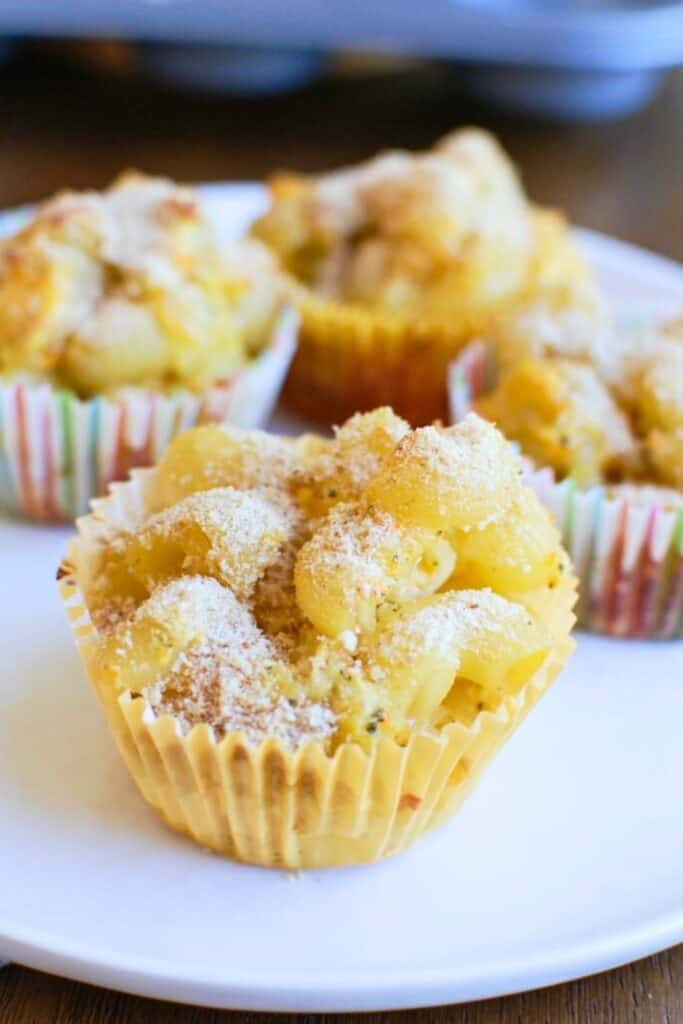 Close-up of a baked mac and cheese muffin in a yellow paper liner, topped with a light dusting of breadcrumbs and Parmesan. The cheesy pasta is formed into a muffin shape for a fun, portable twist on a classic comfort food, with more muffins blurred in the background.