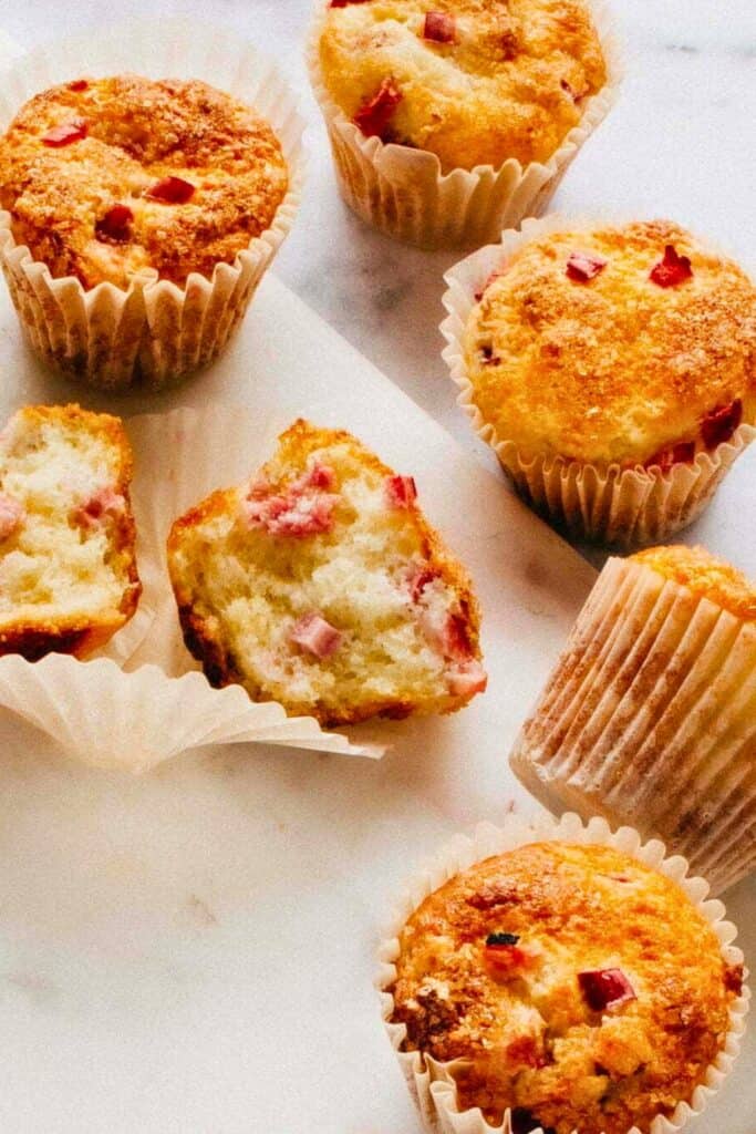 Healthy rhubarb muffins with golden tops and small pink rhubarb pieces baked into a soft fluffy crumb. One muffin is broken open to show the moist interior while others sit scattered on a light surface.