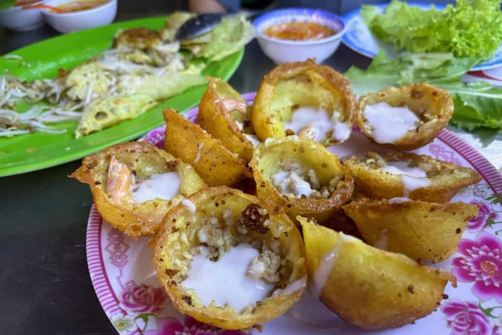 A plate of Vietnamese bánh khọt, small crispy rice flour pancakes topped with shrimp, mung beans, and a drizzle of coconut cream, is served on a floral-patterned dish. In the background, there are leafy greens, dipping sauce, and a partially eaten bánh xèo on a green plate.