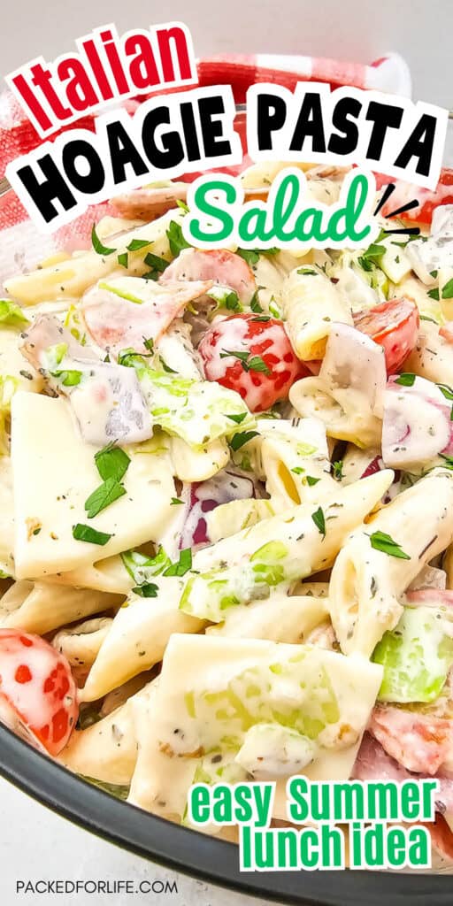 Close-up of a creamy Italian hoagie pasta salad in a glass bowl, featuring penne pasta, lettuce, cherry tomatoes, red onion, sliced deli meats, and provolone cheese, garnished with herbs. Bold text on the image reads 'Italian Hoagie Pasta Salad' and 'easy Summer lunch idea.