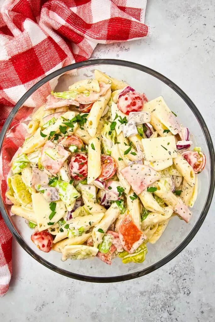 A glass bowl filled with creamy hoagie sub pasta salad, featuring penne pasta, cherry tomatoes, lettuce, sliced deli meats, provolone cheese squares, red onions, and chopped parsley. The bowl rests on a light countertop beside a red and white checkered dish towel, creating a fresh and vibrant picnic-ready vibe.