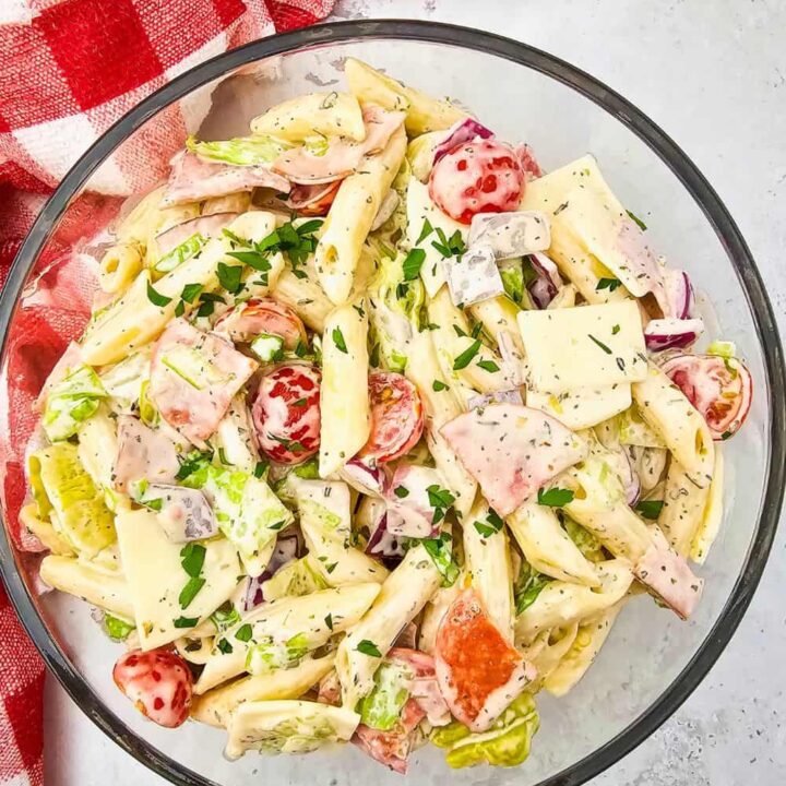 A glass bowl filled with creamy hoagie sub pasta salad, featuring penne pasta, cherry tomatoes, lettuce, sliced deli meats, provolone cheese squares, red onions, and chopped parsley. The bowl rests on a light countertop beside a red and white checkered dish towel, creating a fresh and vibrant picnic-ready vibe.