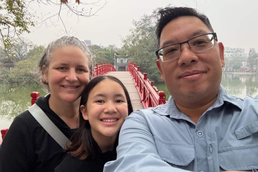 A smiling family poses for a selfie in front of the iconic red bridge leading to Ngoc Son Temple on Hoan Kiem Lake in Hanoi. The peaceful lake and surrounding trees create a calm backdrop, with the temple entrance visible at the end of the bridge.