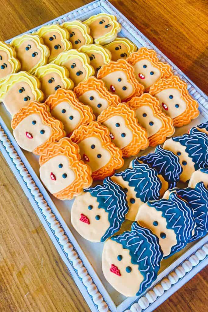 Tray of Sanderson Sisters Hocus Pocus cookies decorated with colorful icing to resemble the three witches from the movie, featuring blonde, orange, and dark blue hairstyles with detailed faces and red lips. The cookies are arranged neatly by character on a white tray over a wooden table.
