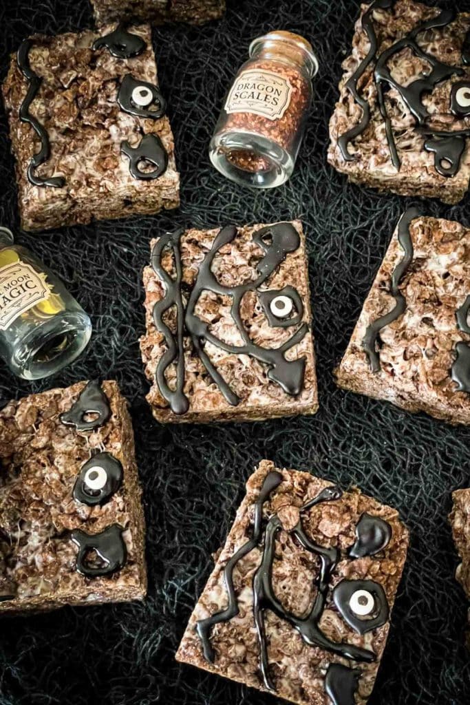 Chocolate Rice Krispies treats decorated as Hocus Pocus spell books with black icing swirls and candy eyes, arranged on a black textured cloth beside small glass jars labeled “Dragon Scales” and “Moon Magic.”