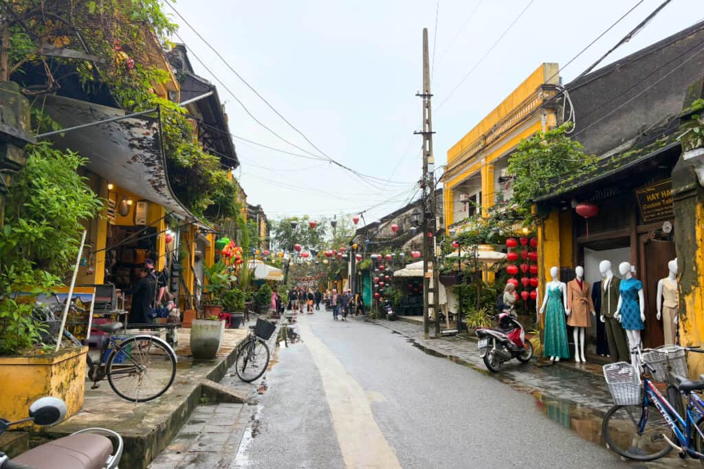 Street in Hoi An Ancient Town lined with yellow buildings, red lanterns, and small shops displaying clothing and souvenirs as people walk along the wet road. Bicycles and motorbikes are parked along the sidewalk, with greenery and hanging lanterns creating a lively historic atmosphere in Hoi An Vietnam.