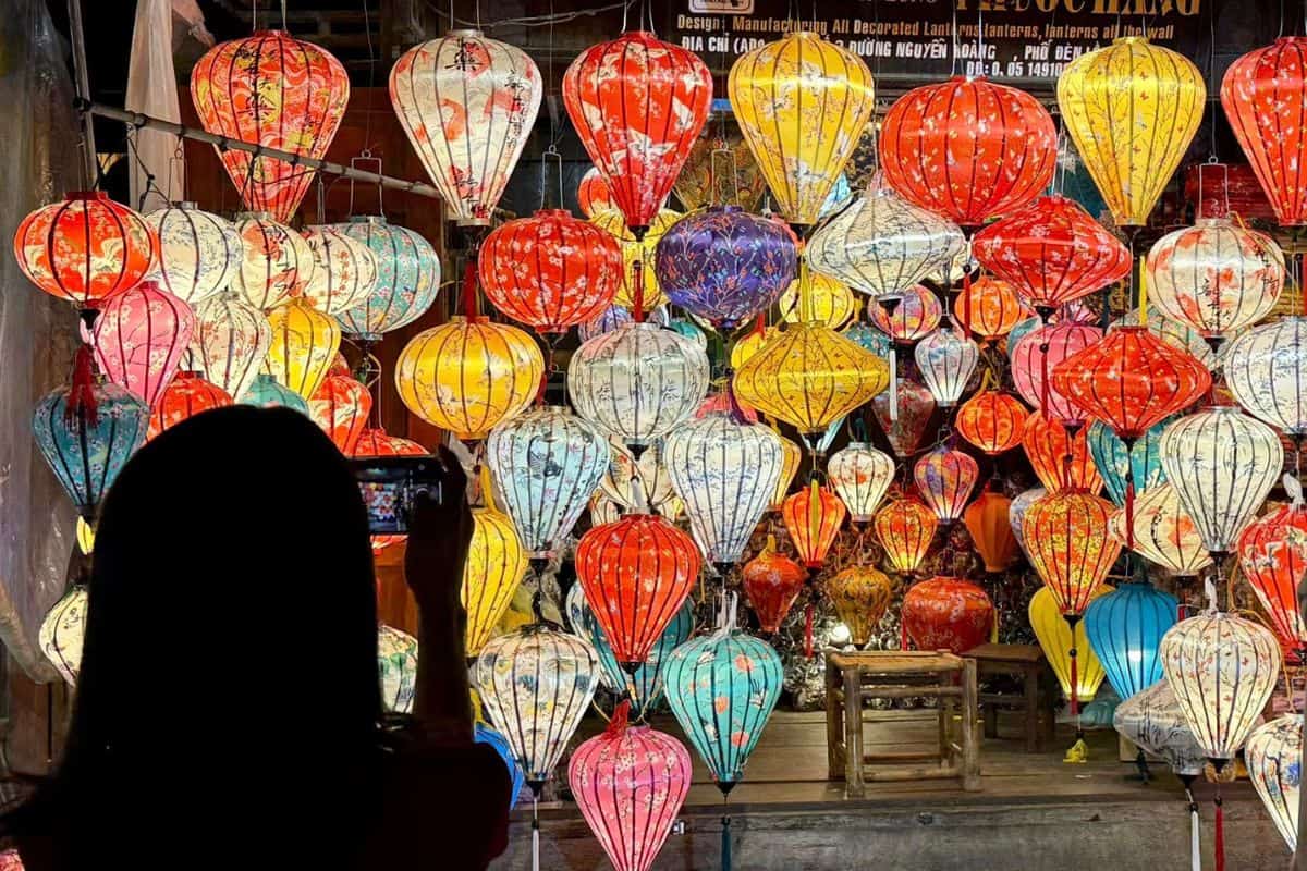 A silhouette of a person holds up a phone to photograph a vibrant display of traditional Vietnamese silk lanterns in various shapes and sizes. The lanterns glow in rich hues of red, yellow, blue, and pink, each decorated with floral and nature-inspired patterns, creating a colorful and festive scene.