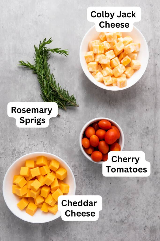 Ingredients for the holiday cheese platter in small and medium bowls; cherry tomatoes, cheddar cheese cubes, colby jack cheese cubes, and sprigs of fresh rosemary.