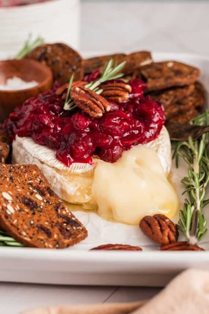 Holiday cranberry baked brie topped with cranberry sauce, toasted pecans, and fresh rosemary, with warm melted cheese spilling from the center. Seeded crackers, extra pecans, and rosemary surround the festive appetizer.
