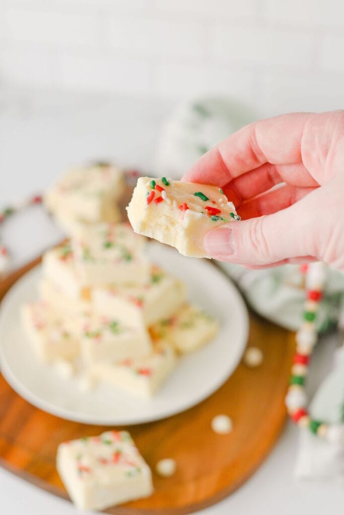Close-up of a hand holding a piece of holiday fudge with a bite taken out, showing the creamy texture inside. The white chocolate fudge is topped with red, green, and white sprinkles, with more pieces stacked on a plate in the festive background.