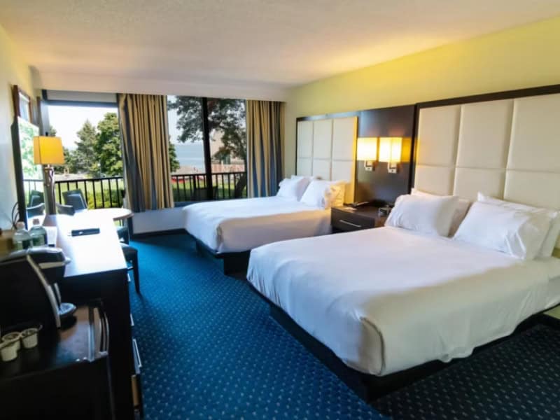 Two queen beds with crisp white linens sit side by side in a spacious hotel room with a blue carpet and modern décor. A sliding glass door opens to a private balcony with a view of trees and distant water, hinting at a peaceful, pet-friendly stay at Holiday Inn resort in Bar Harbor, Maine.
